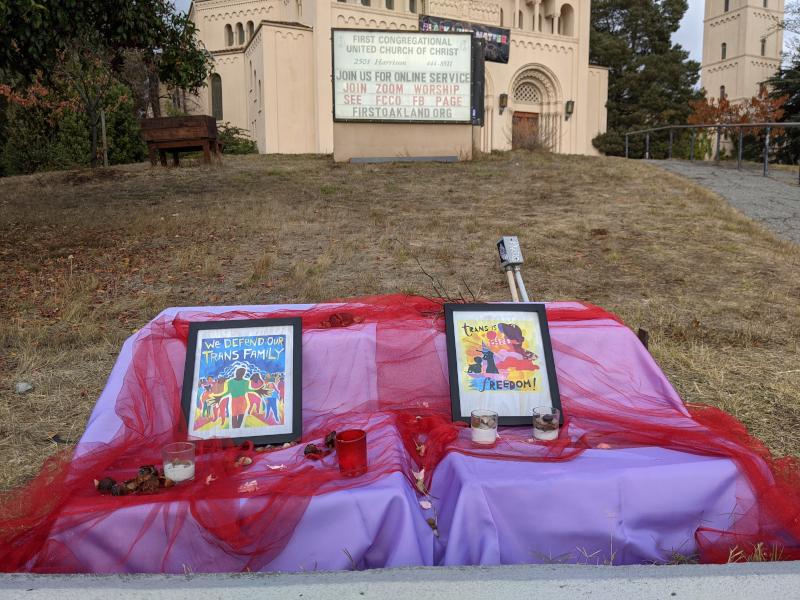 An altar set up in front of a church in Oakland, covered in pink, purple and red fabric. One framed image says 'We Defend our Trans Family,' the other says 'trans is freedom!'